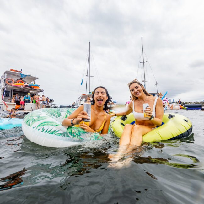 Two women are enjoying a fun day on the water, lounging on inflatable tubes with drinks in hand. Surrounded by boats and other revelers, the atmosphere is lively under clear skies. It’s a social gathering known as The Yacht Social Club Event Boat Charters.