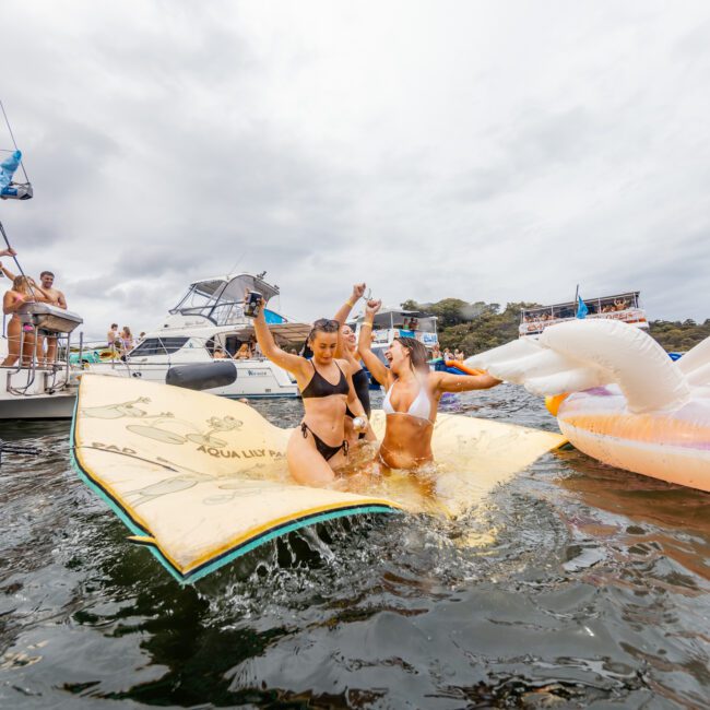 Two people in swimsuits are slipping on a large floating mat on the water, struggling to maintain their balance. Surrounding them are several boats from The Yacht Social Club Sydney Boat Hire, with others cheering from the boats. The sky is overcast.