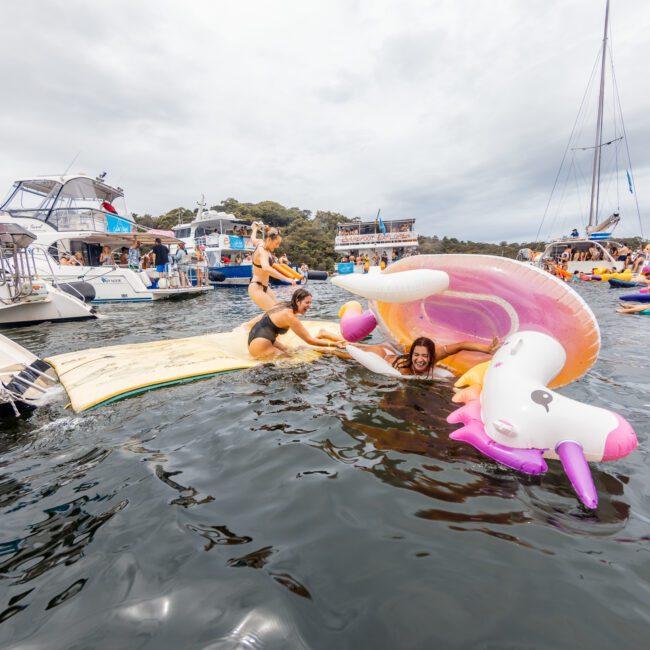 A lively scene on the water features people enjoying colorful inflatable floats, including a giant unicorn. Some are climbing back onto a boat while others float nearby. Numerous boats and yachts from The Yacht Social Club Sydney Boat Hire create a festive atmosphere under an overcast sky.