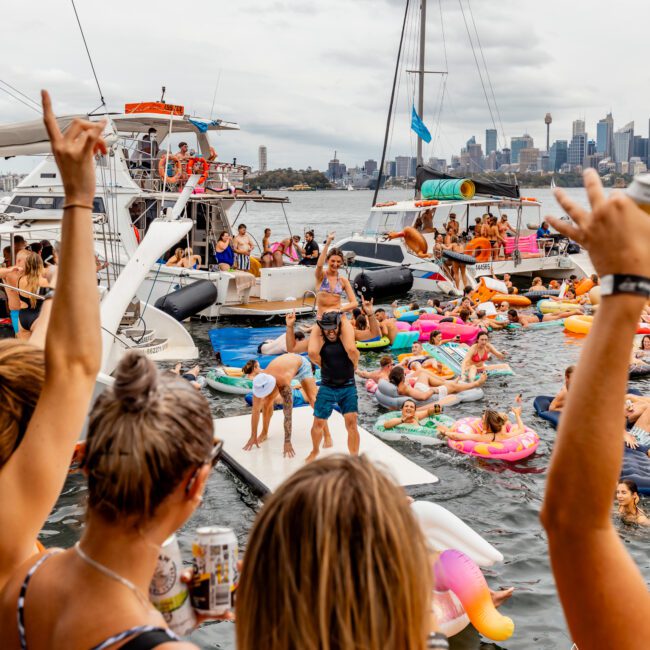 A vibrant scene of a yacht party with numerous people on inflatable floaties in the water. Some guests are dancing on a floating platform, while others cheer and enjoy drinks under the cloudy sky. The Yacht Social Club Sydney Boat Hire provides an unforgettable experience with the city skyline as a backdrop.