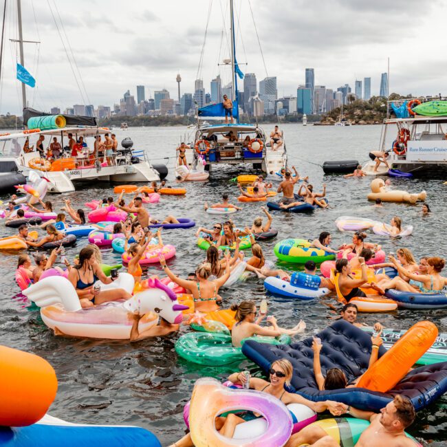 People float on colorful inflatable rafts and pool toys in a busy, lively harbor with several boats nearby, courtesy of Sydney Harbour Boat Hire The Yacht Social Club. A city skyline with tall buildings can be seen in the background under a cloudy sky. The atmosphere is festive and relaxed.