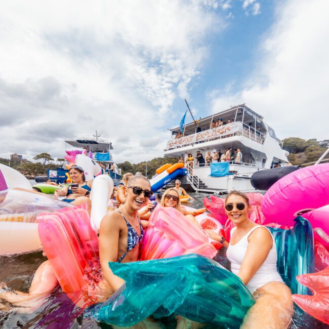 A group of people enjoying a sunny day in the water, surrounded by colorful inflatable floats. Some wear swimsuits and sunglasses, smiling at the camera. A large yacht with a visible "Boat Parties Sydney The Yacht Social Club" banner is anchored in the background.