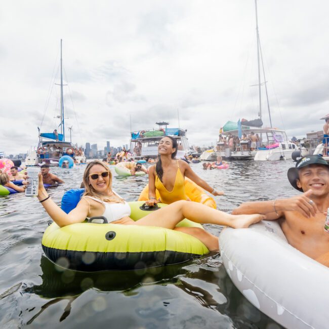 People enjoying the water in inflatable floats on a cloudy day. Boats are anchored nearby, and others in the water are also on inflatables. The mood is festive, with smiles and drinks among the group. A city skyline is visible in the background, exemplifying a classic The Yacht Social Club event with friends.