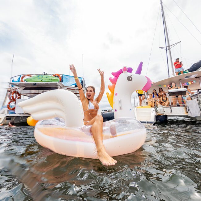 A woman in a white bikini sits on a large inflatable unicorn float in the water, arms raised in joy. Several people are in the background on a sailboat and inflatables, enjoying a sunny day. Experience moments like this with The Yacht Social Club's luxury yacht rentals Sydney.