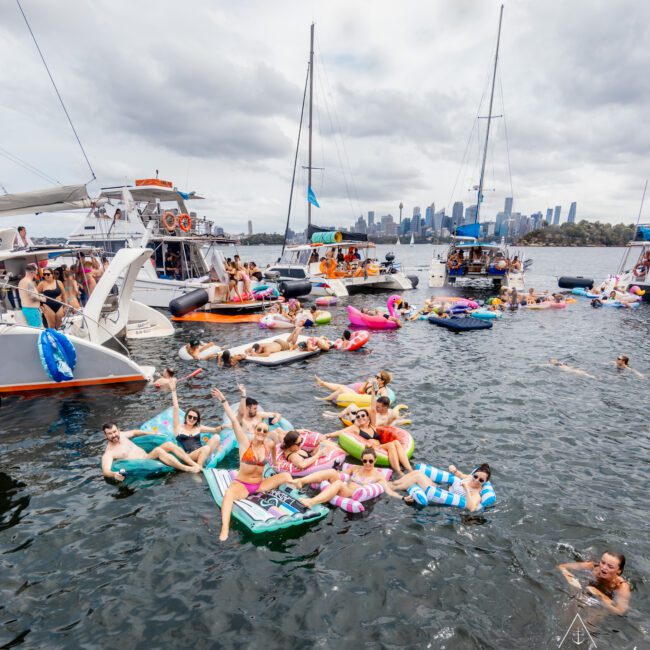 A lively gathering of people on colorful inflatable floats and boats in a body of water. Two large sailboats are anchored nearby, with numerous attendees both on board and in the water. A cityscape with tall buildings is visible in the background under a cloudy sky. Enjoy Boat Parties Sydney The Yacht Social Club!