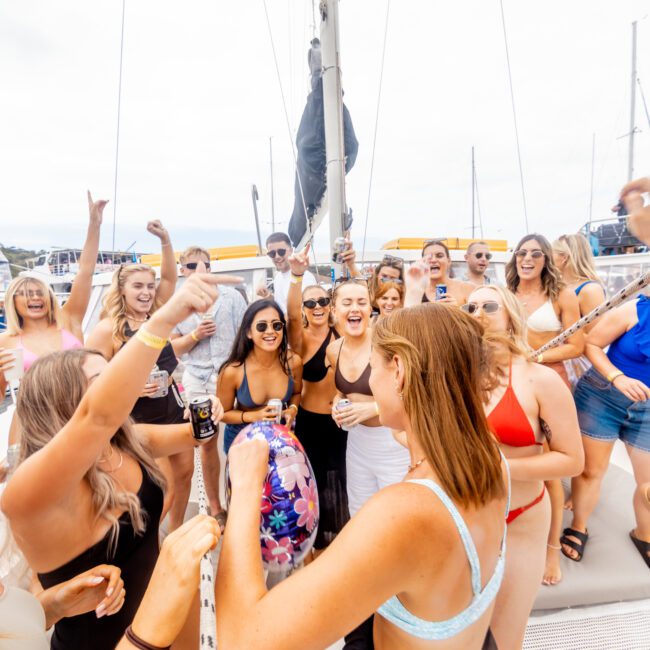 A group of people wearing swimwear and sunglasses are happily celebrating on a sailboat. They are holding drinks, raising their arms in cheer, and smiling. The sky is cloudy, and the boat is docked at a marina with several other boats in the background. Enjoy your experience with Sydney Harbour Boat Hire The Yacht Social Club.