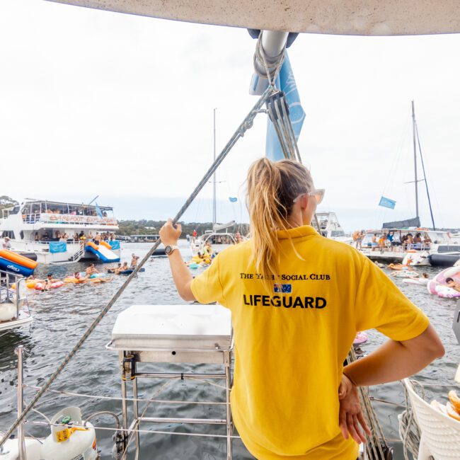 A lifeguard wearing a yellow shirt stands on a boat, surveying a busy scene of people enjoying Boat Parties Sydney The Yacht Social Club on and around various boats and inflatables. Other boats and individuals are visible in the background, soaking in the lively atmosphere.