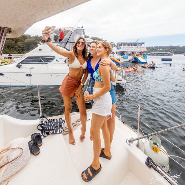 Three women in swimsuits and shorts stand on the deck of a boat, posing for a selfie during The Yacht Social Club Event. Behind them, boats and people in the water are visible, suggesting a lively, festive atmosphere. One woman holds a drink while another raises her arm, holding a hat for the photo.