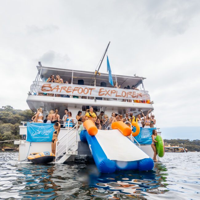 A large boat named "Barefoot Explorer" is anchored, hosting The Yacht Social Club Event. People are enjoying various activities, with a slide extending into the water where several are swimming or floating on inflatables. A banner on the boat reads "Yacht Social." The sky is overcast.