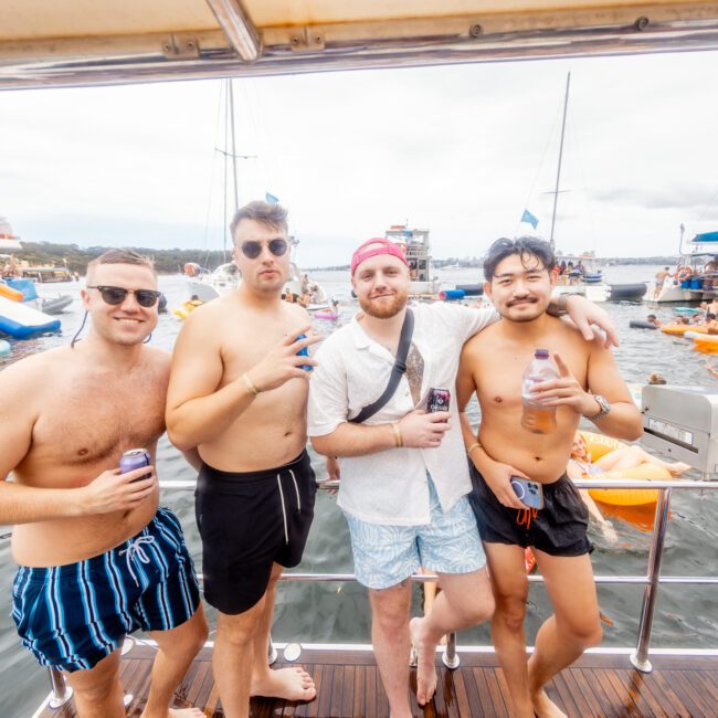 Four men posing on a boat with drinks in hand, smiling at the camera. Dressed in casual summer attire, they enjoy a lively, sunny day on the water. Boats, inflatables, and other people create a festive atmosphere. It looks like an unforgettable event by The Yacht Social Club Sydney Boat Hire.