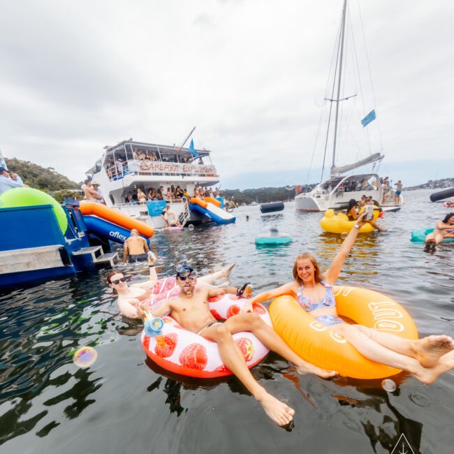 A group of people enjoy a sunny day on the water, lounging on inflatable pool floats. In the background, boats with additional revelers are anchored, creating a lively party atmosphere. The scene includes water toys and promotes a festive vibe typical of Sydney Harbour Boat Hire by The Yacht Social Club.