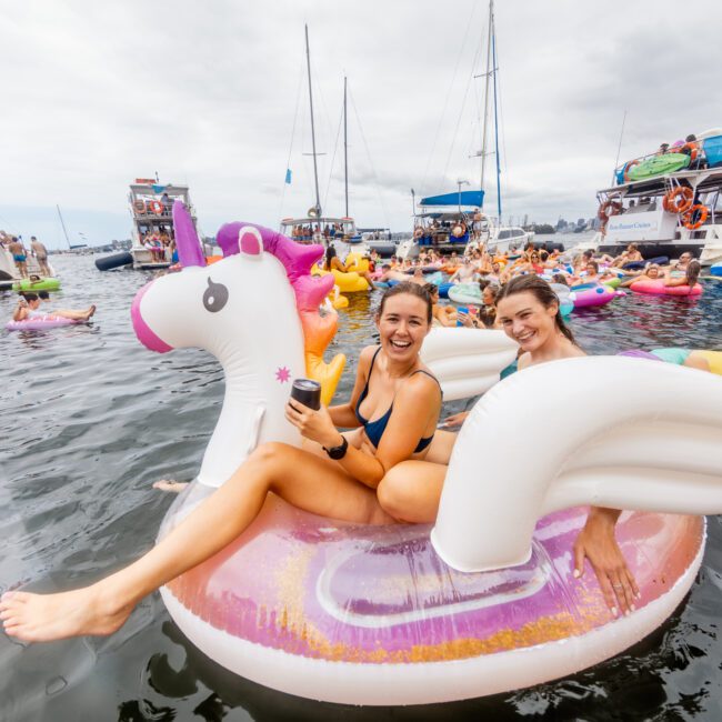 Two women in swimsuits smile and sit on a large inflatable unicorn float in the middle of a lake, surrounded by other people on various inflatables and boats. The scene is festive and lively, with cloudy skies above. Text in the bottom corner reads "Boat Parties Sydney The Yacht Social Club.