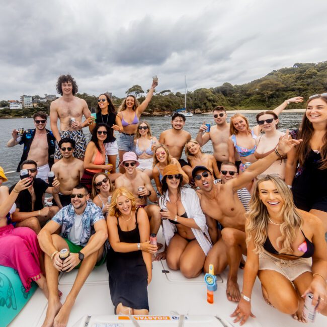 A large group of people is gathered on a boat, smiling and posing for the camera. They are dressed in swimsuits and casual summer attire, enjoying a day out on the water with The Yacht Social Club. Trees and a cloudy sky serve as the backdrop.