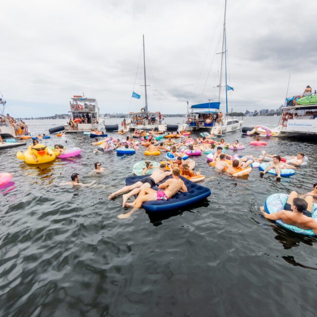 A group of people are relaxing and socializing on colorful inflatable floats in a crowded lake, surrounded by several anchored boats on a cloudy day. Some people are sunbathing, while others are chatting or enjoying the water. Flags wave above the luxury yachts from The Yacht Social Club Sydney Boat Hire.