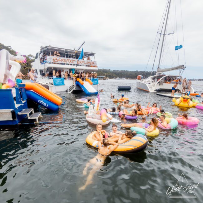 A group of people enjoying a festive gathering in a lake, with many floating on colorful inflatables near a large boat named "Barefoot Explorer." More people are seen on a yacht nearby, showcasing The Yacht Social Club Sydney Boat Hire. The event appears to have a lively and fun atmosphere.