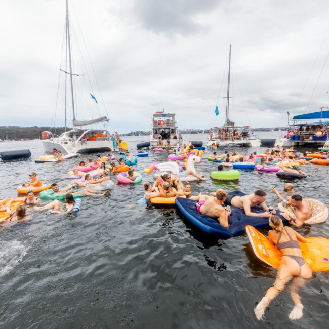 People enjoying a lively boat party on the water, surrounded by yachts and various inflatable floats. The attendees are swimming, floating, and socializing under a cloudy sky, creating a festive atmosphere at The Yacht Social Club Sydney Boat Hire.