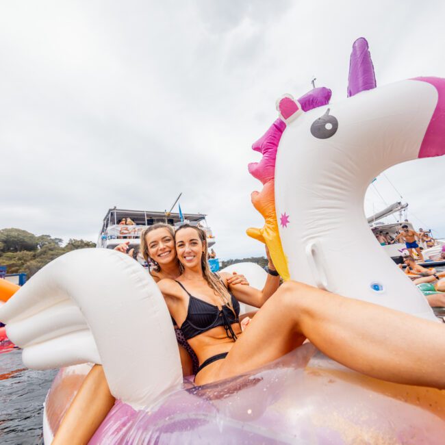 Two women in swimsuits are enjoying themselves on an inflatable unicorn float in the water, smiling at the camera. In the background, other people are seen on a yacht and additional inflatable floats. The atmosphere is lively and festive, reminiscent of a fun day with The Yacht Social Club Event Boat Charters.