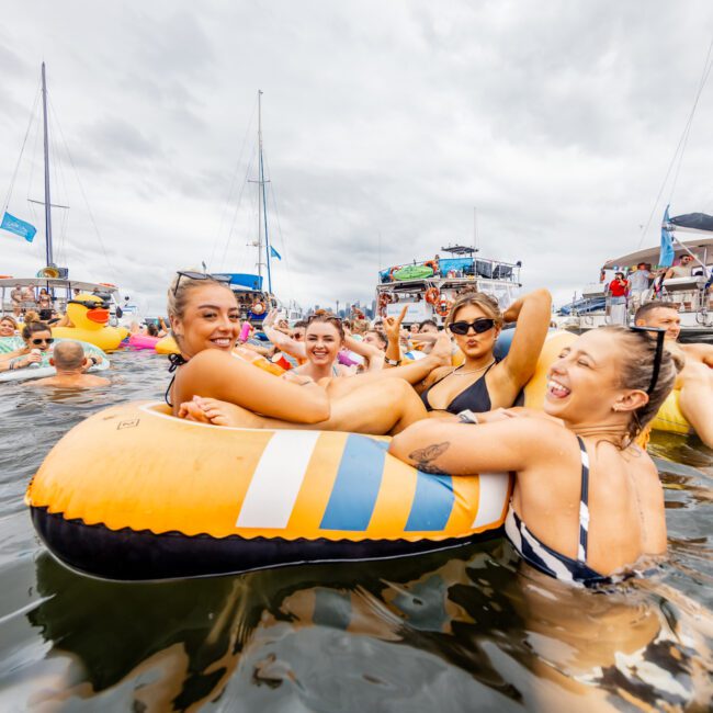 A group of people happily floating in a large, colorful inflatable raft on the water among several boats. The sky is cloudy, and the atmosphere is lively with many others lounging on inflatables or yachts in the background. Perfect for a day with Sydney Harbour Boat Hire The Yacht Social Club.