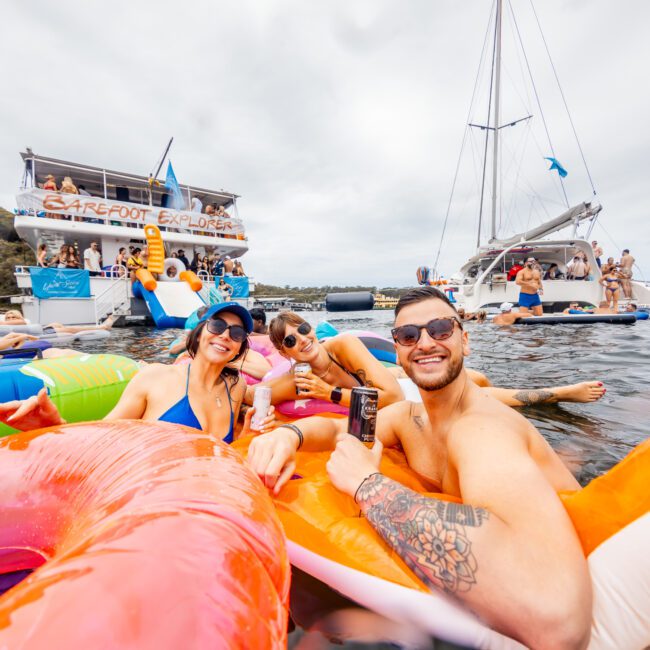 A group of three people smiling and holding drinks while floating on inflatable tubes in the water. Behind them, other people are on boats, including a large boat with "Yacht Social Club" written on it. Experience the fun with Luxury Yacht Rentals Sydney for an unforgettable day. The sky is overcast.