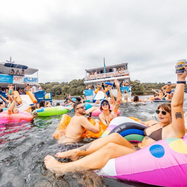 A lively group of people enjoy a party on the lake, floating on colorful inflatables. Two boats, including one with "The Yacht Social Club Sydney Boat Hire" branding, are in the background. The atmosphere is festive, with many people holding drinks and cheering.