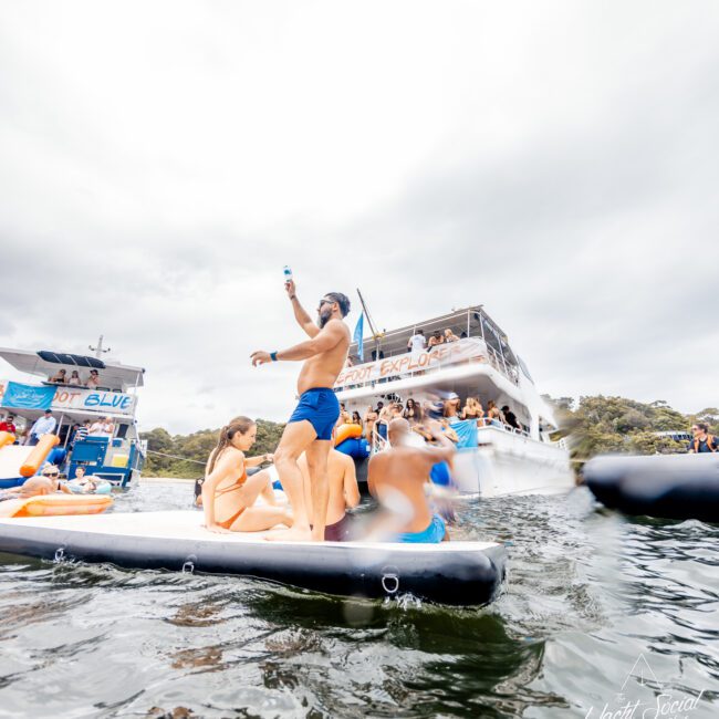 A group of people in swimsuits celebrate on an inflatable platform in the water near two double-decker party boats. The sky is overcast, and trees are visible in the background. Some are sitting, while others stand and dance, enjoying a lively Boat Parties Sydney The Yacht Social Club event.