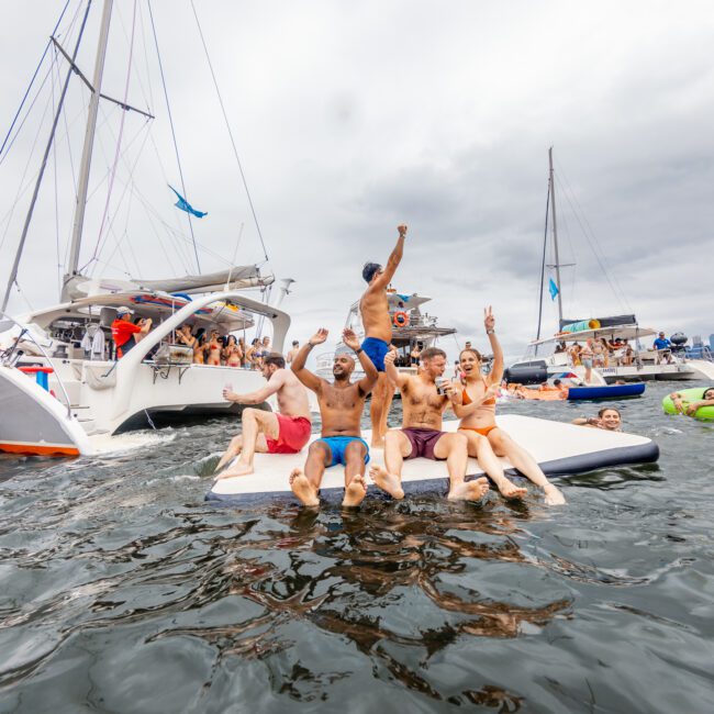 A group of people are enjoying themselves on floating foam mats in the water near several yachts. Some are raising their arms in celebration, and others are swimming nearby. The sky is overcast, and the atmosphere is lively and festive. The text "The Yacht Social Club Event Boat Charters" is visible in the bottom right corner.