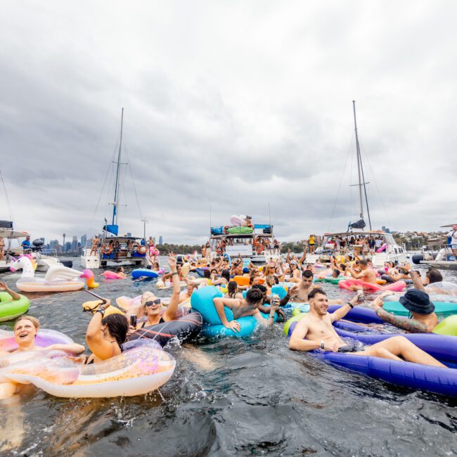 A lively scene at a boat party on the water, with many people enjoying the event on colorful inflatables, surrounded by multiple boats. The atmosphere is vibrant and festive despite the overcast sky. Hosted by The Yacht Social Club, Sydney Harbour's skyline adds a stunning backdrop to this unforgettable experience.