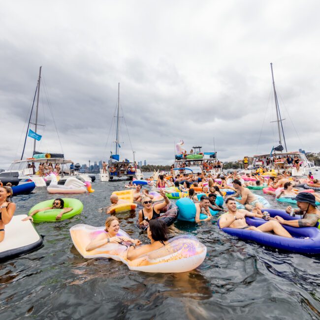 A lively scene of people enjoying a Yacht Social Club Event in a marina. The attendees lounge on colorful floaties in the water while others dance and socialize on boats from Luxury Yacht Rentals Sydney. The sky is overcast, creating a contrast with the vibrant atmosphere.