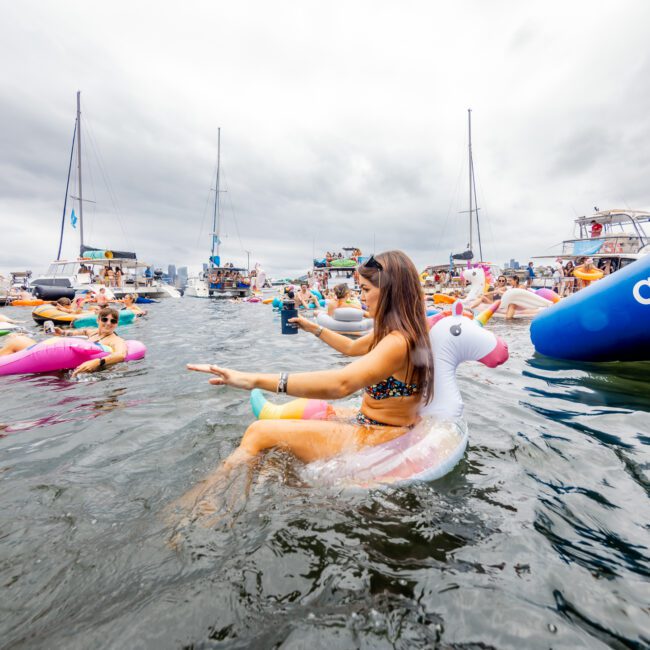 A vibrant scene of people in swimsuits enjoying a boat party on the water with colorful inflatable floats. One woman is centered, sitting on a unicorn float while holding a drink and smiling. Boats and other floats from The Yacht Social Club Sydney Boat Hire are visible in the background against an overcast sky.