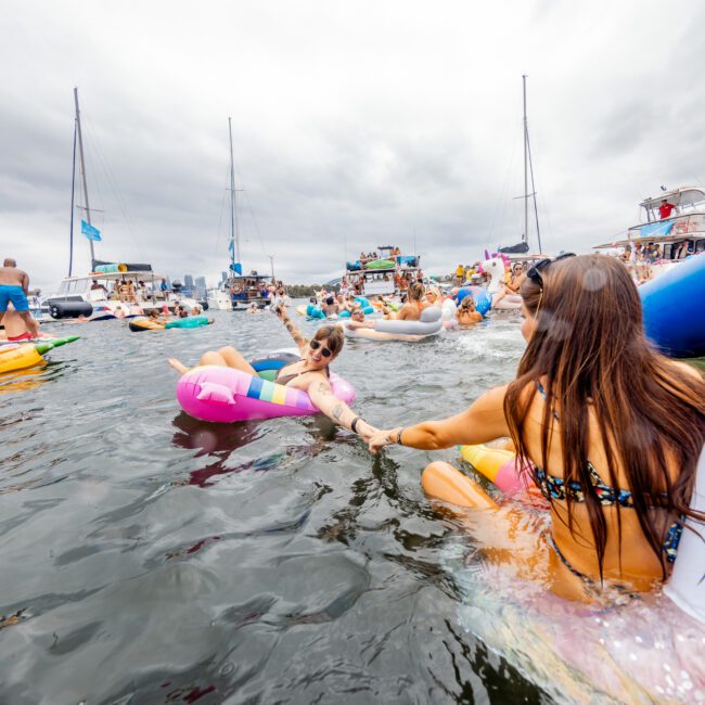 A lively lake event with numerous people on inflatable floats of various shapes and colors, including a unicorn float. Boats from The Yacht Social Club can be seen in the background under a cloudy sky. Two women in swimsuits hold hands while floating on the water.