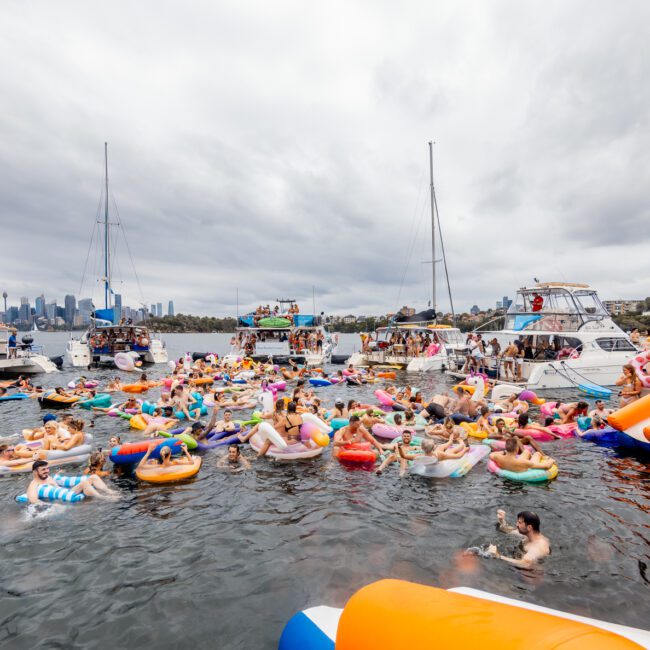 A lively scene of a floating party with numerous people on colorful inflatable floats in the water. Several boats are anchored around, highlighting The Yacht Social Club Sydney Boat Hire. The skyline of a city is visible in the background under a cloudy sky. A blue and orange inflatable slide is on the right side.