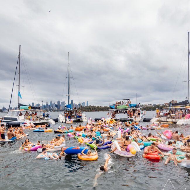 A large group of people are enjoying a festive party on the water, floating on colorful inflatable rafts and tubes. In the background, there are several boats anchored and a city skyline visible under a cloudy sky. The Yacht Social Club Sydney Boat Hire adds to the lively scene with its elegant vessels.