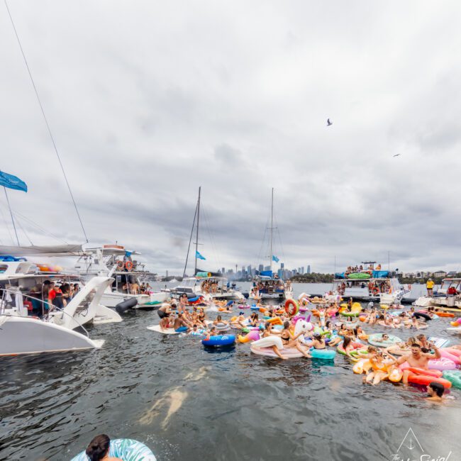 The Yacht Social Club's lively gathering sees people enjoying a sunny day on water floats, inflatables, and boats. The background shows more boats and a distant city skyline. The event appears festive, with individuals swimming and relaxing on colorful floaties in Sydney Harbour.