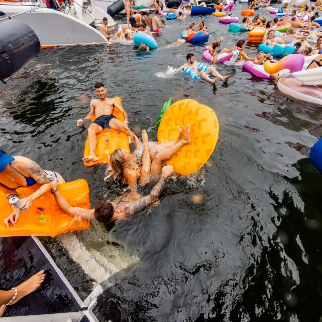 People enjoy a party on a lake, lounging on inflatable floats in the water near several docked boats. Some are swimming or floating, while others socialize on the boats. The mood appears lively and festive, akin to an event hosted by Luxury Yacht Rentals Sydney.