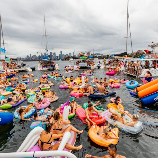 A vibrant scene on the water with numerous people enjoying a floating party by Boat Parties Sydney The Yacht Social Club. Various inflatable rafts and floaties with colorful designs cover the water surface. Boats are docked in the background, and the sky is overcast with a city skyline visible.