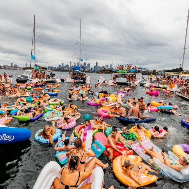 A large group of people lounging on colorful inflatable floats in the water, with several boats in the background and a city skyline in the distance. The scene appears joyful and festive under cloudy skies, reminiscent of Boat Parties Sydney The Yacht Social Club events.