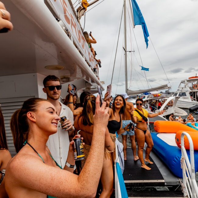 A group of people in swimwear enjoying a lively Boat Parties Sydney The Yacht Social Club event on a boat. Some are taking selfies, while others hold drinks. The deck is bustling, with inflated pool floats and a slide visible in the water. The sky is cloudy, indicating an overcast day.