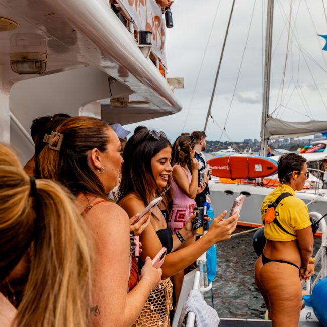 A group of people on the deck of a boat, taking photos and enjoying themselves at The Yacht Social Club Event. Some are holding drinks and one person is wearing a yellow life jacket. Other boats and the ocean are visible in the background. The atmosphere appears festive and social.