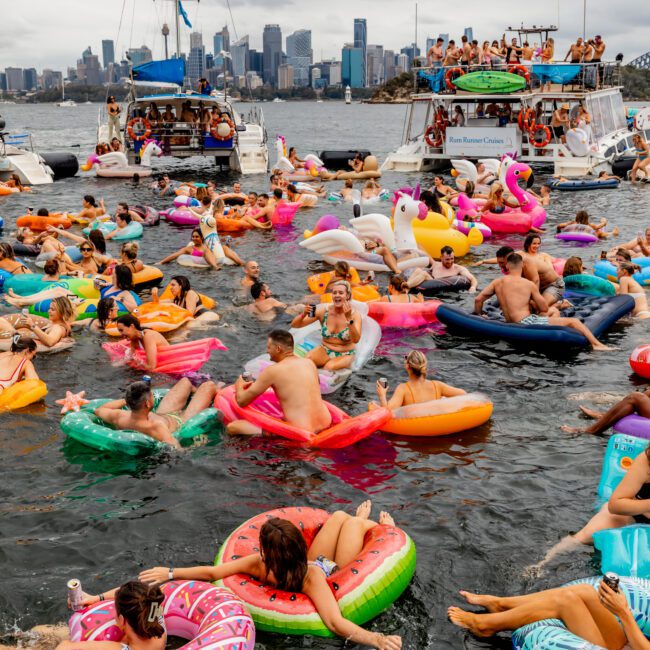 A lively scene of people on colorful inflatable floats in a body of water, surrounded by boats. The sky is overcast, and a city skyline is visible in the background. People are socializing and having fun amidst vibrant floats at The Yacht Social Club Event Boat Charters.