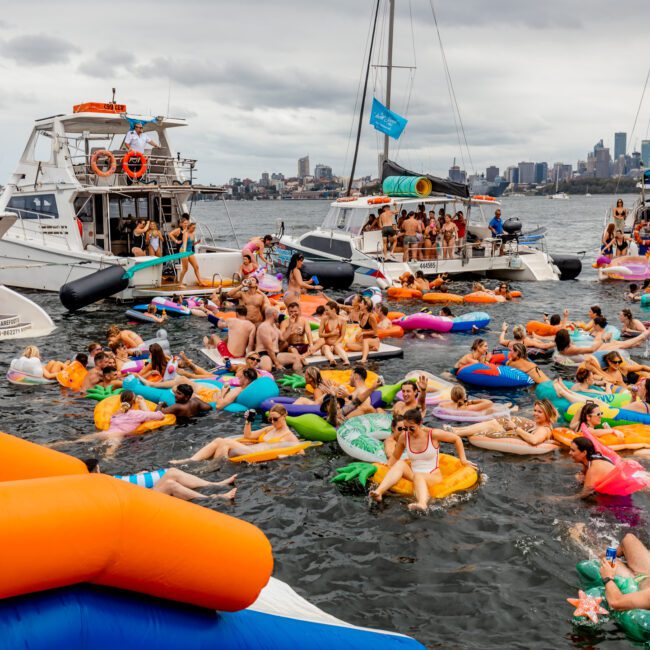 A lively scene of a boat party on water, featuring numerous people on colorful inflatables and floating toys. Several yachts and boats from The Yacht Social Club Sydney Boat Hire are moored closely together, while a city skyline is visible in the background under a cloudy sky.