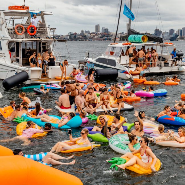 A lively group of people relaxing on colorful inflatable floats in the water, surrounded by boats on a cloudy day. Many are enjoying drinks and laughter at The Yacht Social Club Event Boat Charters. In the background, a cityscape is partially visible.