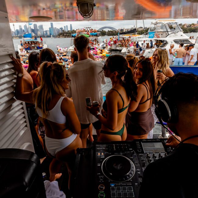 A DJ is performing on a boat with a mixing console, while people in swimwear dance and socialize. A large group gathers on another party boat nearby. The city skyline is visible across the water, adding to the atmosphere of The Yacht Social Club Sydney Boat Hire event.