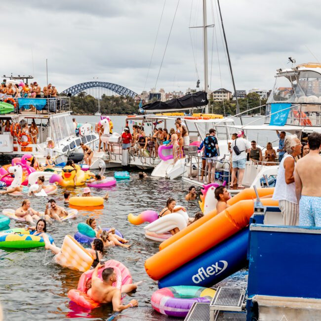 A lively scene of people enjoying a yacht party on the water. Several yachts are docked close together, with partygoers swimming and floating on colorful inflatables. The Yacht Social Club Sydney Boat Hire offers unforgettable experiences, with the backdrop featuring a cloudy sky and a bridge in the distance.