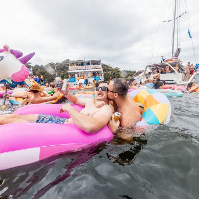 A group of people are seen enjoying a party on a lake. Two people are sitting on a large pink float with a unicorn head. Other colorful inflatables and boats from Sydney Harbour Boat Hire are in the background. The atmosphere is lively and festive, with everyone smiling and having fun.