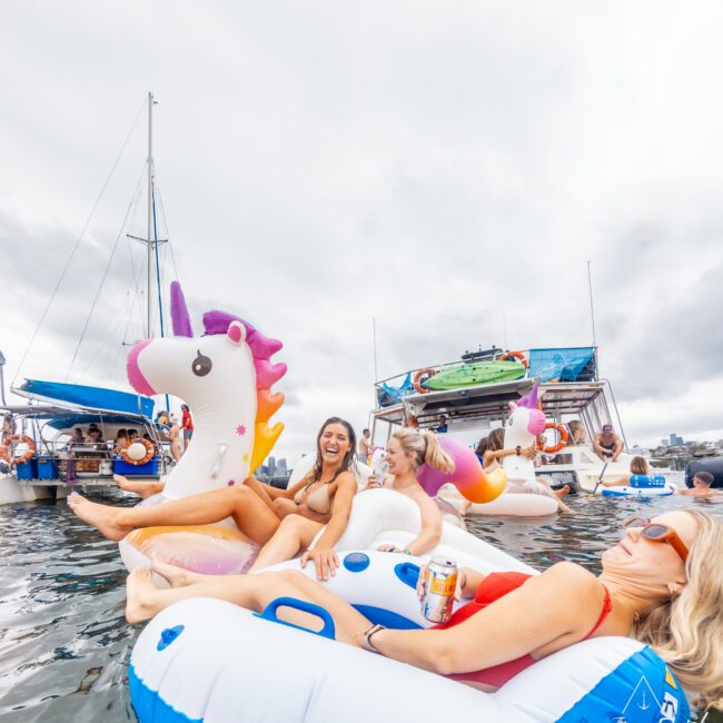 Three women relax on inflatable unicorn and circle floaties in the water, smiling and enjoying themselves. In the background, boats and other people are visible, creating a festive atmosphere under a cloudy sky. The Yacht Social Club Event Boat Charters logo is visible in the bottom right corner.