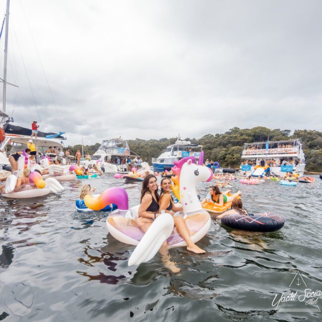 A crowded water scene features numerous people on inflatable floats, including a unicorn, cupcake, and doughnut. Two women, smiling and wearing swimsuits, sit on the unicorn float. Boats from The Yacht Social Club Event Boat Charters are docked nearby with a forested shoreline in the background.