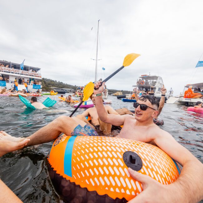 A group of people enjoying a day on the water, floating on inflatable tubes and paddling with oars. Boats and other inflatables are visible in the background, along with people swimming and relaxing. The atmosphere is lively and festive, reminiscent of a gathering with Sydney Harbour Boat Hire The Yacht Social Club.