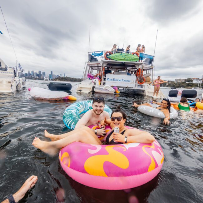 A group of people is enjoying a day on the water near a large boat. Some individuals are lounging on colorful inflatables under an overcast sky. In the background, more people are on the boat, smiling and socializing at The Yacht Social Club Event Boat Charters in Sydney Harbour.