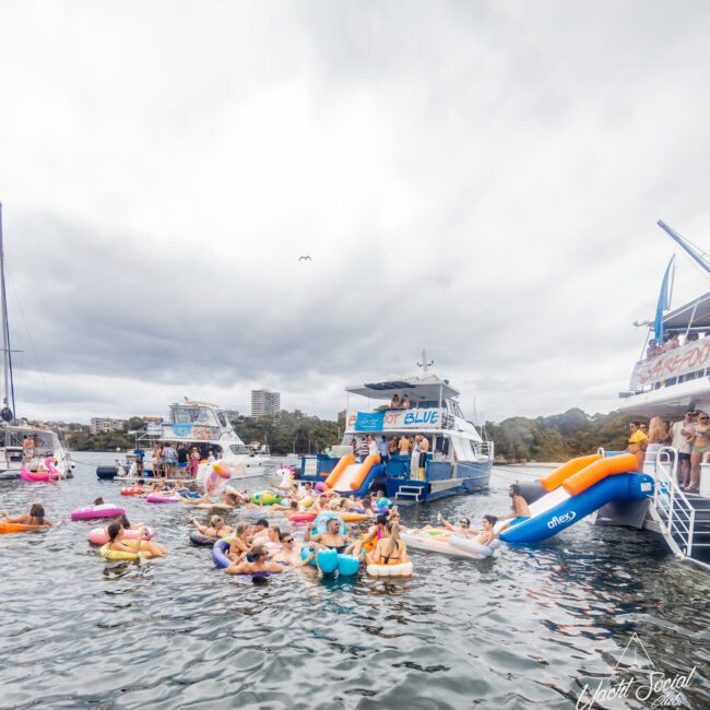 A lively scene of people enjoying a yacht party on a cloudy day. Several yachts, part of Boat Parties Sydney The Yacht Social Club, are anchored close to each other, with many people swimming and floating on colorful inflatables. Inflatable slides are mounted on some yachts, adding to the fun atmosphere.