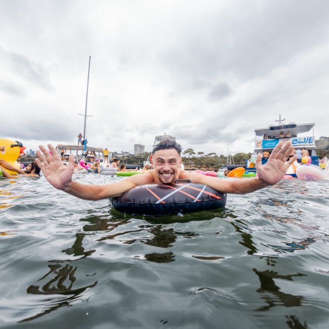 A man smiles and raises his arms while floating on an inflatable device in a lake, surrounded by other people on similar inflatables. The sky is overcast, and buildings and trees are visible in the background. It’s reminiscent of the relaxed vibes of a Boat Party with The Yacht Social Club in Sydney.